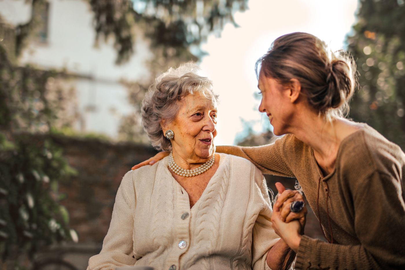 An adult daughter holding hands with her smiling elderly mother in a sunlit garden